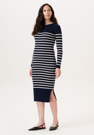 Femme debout portant une robe en maille à manches longues rayée horizontale bleu marine et blanche, avec une fente sur le côté et des chaussures plates noires à bout pointu.
