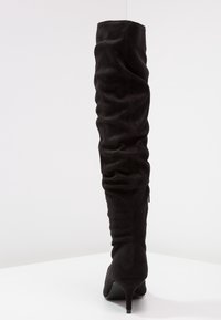 Bota negra por encima de la rodilla en suave ante con un diseño holgado, punta afilada y un tacón delgado. Cuenta con una cremallera en el lateral.