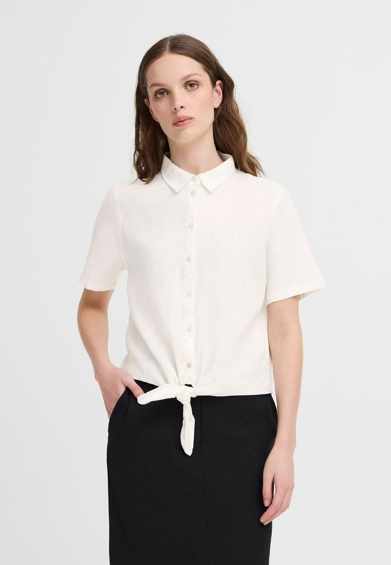 Camicia bianca in lino a maniche corte con chiusura a bottoni e dettaglio annodato in vita, abbinata a una gonna nera.