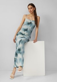 Lichtblauwe maxi-jurk met een tie-dye patroon, dunne bandjes, aangesloten silhouette en een gladde texture. Gecombineerd met witte sandalen met hakken en banden.