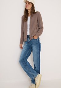 Lichtbruine gebreide cardigan met ritssluiting, witte t-shirt, blauwe jeans met contrasterende zijpaneel en beige sneakers.