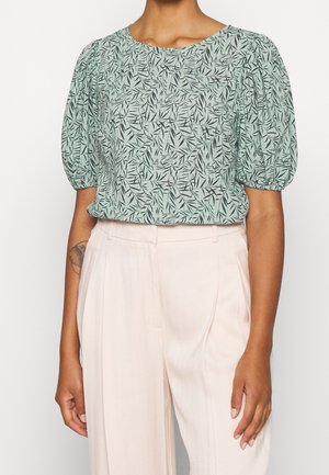 Woman wearing light green leaf-patterned puff sleeve blouse tucked into high-waist cream pleated pants, arm tattoo visible.