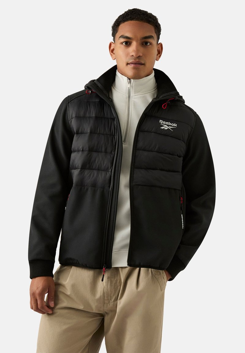 Pikowana czarna kurtka z kapturem, białymi akcentami przeszyć, kieszeniami na zamek i logo na piersi. Noszona na jasnoszarym topie.