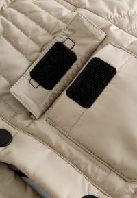 Nærbillede af beige quiltet stof med to sorte Velcro-lukninger syet på rektangulære klapper for sikker lukning.