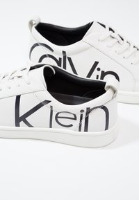 Paire de baskets blanches basses avec intérieur noir et grandes lettres noires imprimées sur les côtés sur fond blanc.