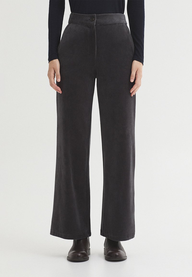 Femme portant un pantalon large en velours côtelé gris foncé taille haute avec boutons sur le devant et des bottines noires sur fond blanc.
