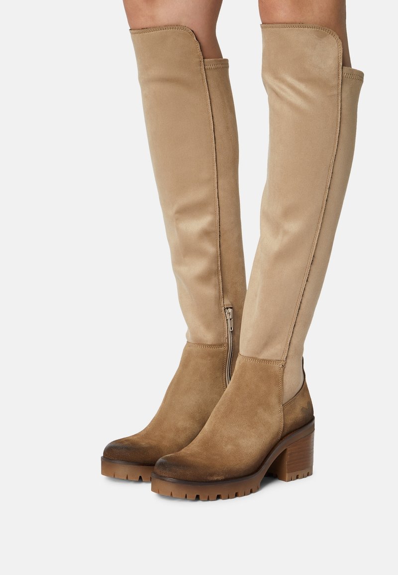 Bottes en daim beige montant jusqu'au genou avec talons carrés, fermetures éclair latérales et semelles à crampons portées sur des jambes sur un fond uni.