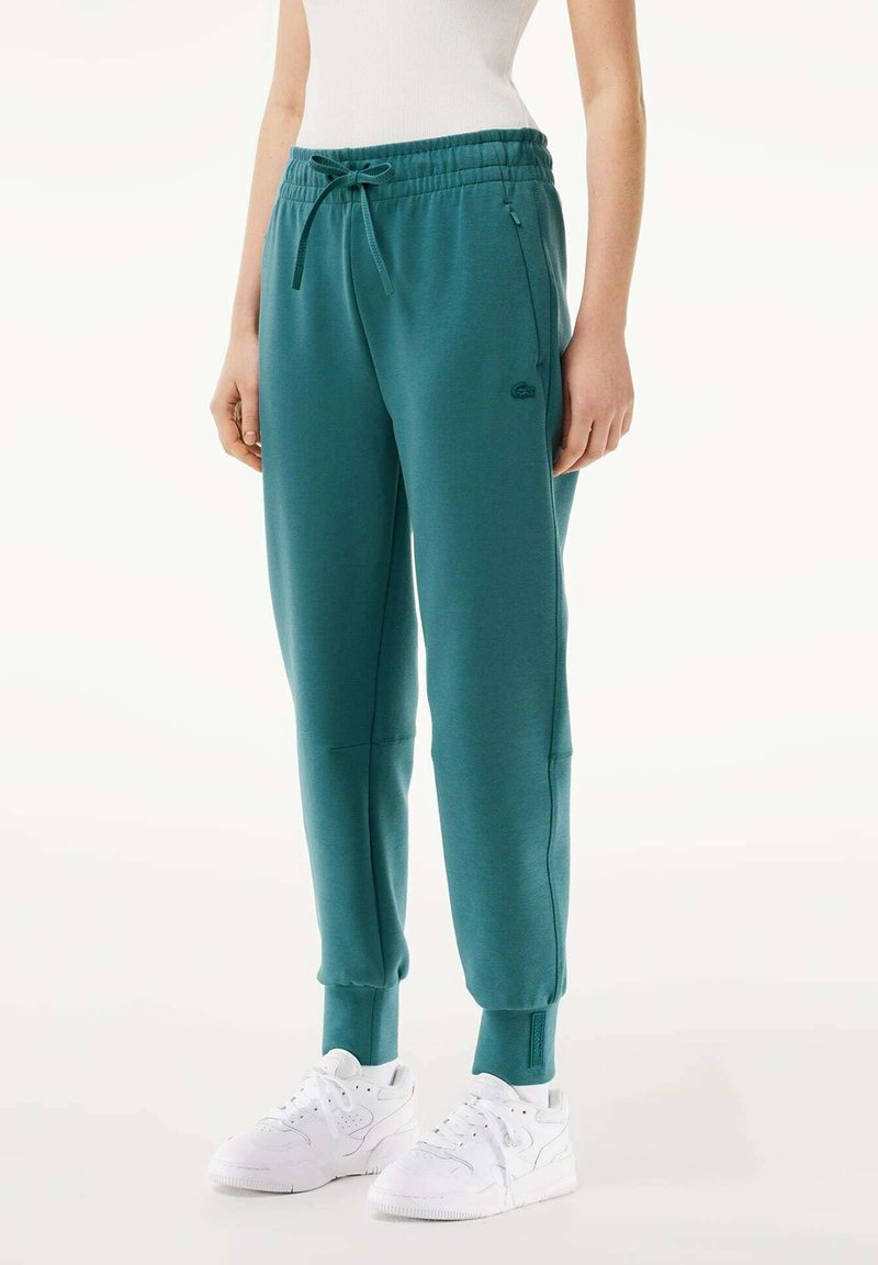 Turkusowe dresy wykonane z miękkiego materiału, z elastycznym paskiem w talii i sznurkiem, bocznymi kieszeniami oraz ściągaczami na końcach nogawek. W zestawie z białymi trampkami.