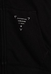 Haut à zip noir avec une texture douce, une petite poche frontale dotée d'un logo graphique blanc, et des accents discrets en blanc.
