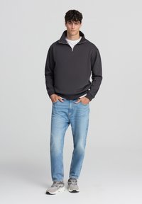 Felpa con zip grigia scura, t-shirt bianca a girocollo sotto, abbinata a jeans azzurri chiari e sneakers grigie. Look casual, design minimale.