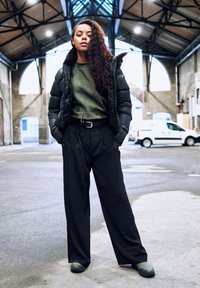 Gepolsterte schwarze Jacke, grünes geripptes Shirt, hochgeschnittene schwarze Hose und dunkelgrüne Slipper, in einem industriellen Raum mit gewölbten Fenstern.
