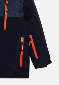 Marinejakke med teksturert mørkeblå yoke, oransje glidelåser og flere lommer. Har et quiltesdesign og en tettsittende mansjettåpning.