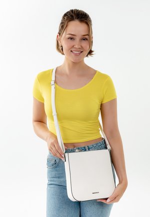 Bílá kožená crossbody taška s černým obšitím, obdélníkový tvar, nastavitelný popruh a texturovaný povrch, kterou drží osoba v žlutém topu.
