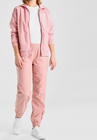 Light pink zip-up jacket with ribbed cuffs and elastic waistband, paired with matching lightweight pants. White T-shirt and sneakers.