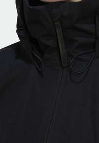 Veste à capuche noire avec fermeture éclair à l'avant et cordons réglables au niveau du col, couvrant la zone du cou et du menton.