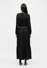 Μαύρο μάξι φόρεμα με υφή υφάσματος, μακριά μανίκια και σχεδιασμό φούστας με στρώσεις. Διαθέτει ντεκολτέ με βολάν και μεταλλική ζώνη αλυσίδα.