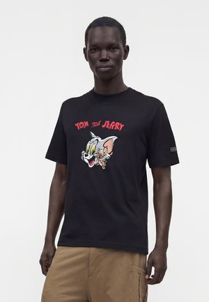 Homme portant un T-shirt noir avec des visages de dessin animé Tom et Jerry et un texte rouge, associé à un pantalon cargo beige, debout devant un fond uni.