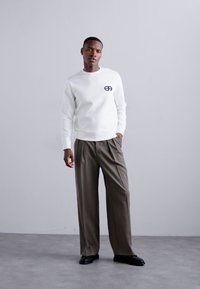 Sweatshirt branco com um pequeno logotipo preto no peito, combinado com calças largas de tom castanho-claro e sapatos pretos elegantes.