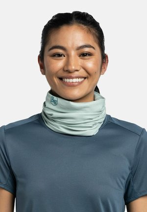 Young woman smiling, wearing a light green Buff neck gaiter and a dark blue short-sleeve athletic shirt, plain light background.