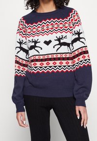 Marinblå tröja med röda och vita geometriska mönster, prydd med svarta renar och ett hjärtmotiv. Ribbad manschett och fåll, stickad struktur.