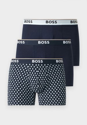 Muskulózny muž stojaci bosý so založenými rukami, oblečený v tmavomodrých boxerkoch s logom „BOSS“ na páse, na jednofarebnom pozadí.