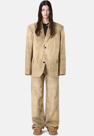 Personne aux longs cheveux foncés porte un costume beige à motifs surdimensionné avec pantalon large et chaussures assorties, debout devant un fond uni.