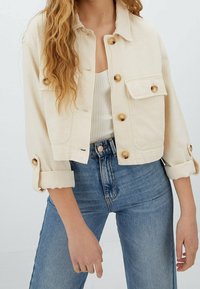 Giacca in denim beige corta con colletto, chiusura a bottoni, tasca sul petto e maniche arrotolate. Tessuto strutturato con dettagli delle cuciture visibili.