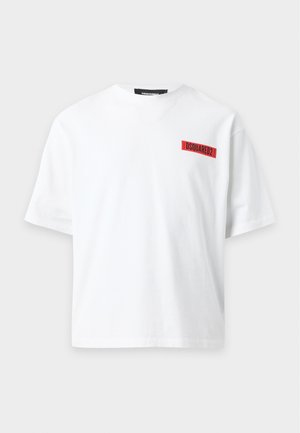 Joven con camiseta blanca con logo rojo, vaqueros azules, zapatillas blancas, collares superpuestos y cinturón negro con hebilla de hoja de arce roja.