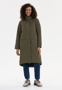 Olijfgroene lange jas met een hoge kraag, voorzien van een ritssluiting aan de voorkant, twee voorzakken en koordaccenten. Gedragen over blauwe spijkerbroeken en sneakers.