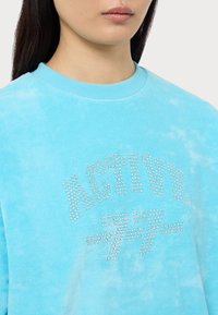 Sudadera azul claro con una textura suave, que presenta un gráfico de "ACTIVE" en brillantes de strass en el pecho y un diseño de cuello redondo.
