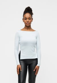 Ανοιχτό μπλε μακρυμάνικη μπλούζα με λαιμόκοψη σκάφος, απαλό ύφασμα και ρυτιδωμένη λεπτομέρεια στο πλάι, σε συνδυασμό με μαύρο εφαρμοστό κολάν.