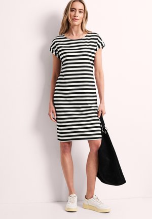 Kobieta w czarno-białej, poziomo paskowanej sukience do kolan, białe trampki, trzyma dużą czarną torbę na jednolitym tle.