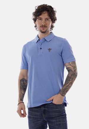 Uomo con capelli ricci e tatuaggi su entrambe le braccia, indossa una polo azzurro chiaro e jeans scuri, in piedi con una mano in tasca.