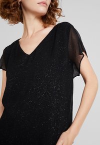 Black top with V-neck, short sheer sleeves, and textured fabric featuring metallic flecks throughout for added detail.