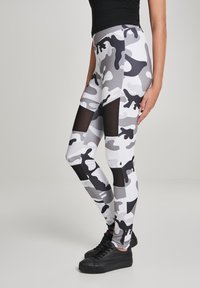 Szürke álcázós leggings, átlátszó hálós panelek a lábak mentén. Rugalmas anyagból készült, testhezálló fazon, fekete sportcipővel párosítva.