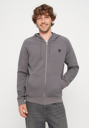 Jeune homme aux cheveux bouclés portant un sweat à capuche gris zippé avec des poches et des cordons, souriant avec une main dans la poche.