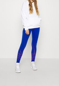 Leggings de sport bleus avec des accents de lignes roses, associés à un sweat à capuche blanc. Le matériau semble extensible et lisse. Associé à des baskets blanches.