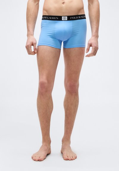 Άνδρας που φοράει ανοιχτό μπλε μποξέρ με μαύρη μέση και την ετικέτα "Polo Ralph Lauren" στέκεται ξυπόλητος σε μονόχρωμο γκρι φόντο.