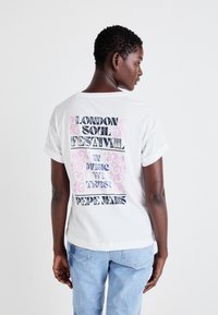 Balts kokvilnas T-krekls ar grafisku druku uz muguras, kurā ir uzraksts "LONDON SOUL FESTIVAL" melnā un rozā dekoratīvā tekstā. Īsas piedurknes.