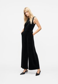 Femme blonde debout, les mains dans les poches, portant une combinaison noire sans manches à jambes larges et des escarpins noirs à bout pointu avec bride arrière, sur fond blanc.