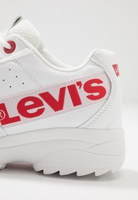 White sneaker with a red "Levi's" logo on the side, featuring a smooth leather upper, textured heel, and rubber outsole with a wave pattern.