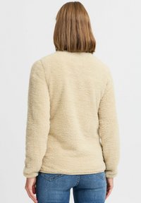 Beige fleece pullover with a soft, textured exterior, long sleeves, and a round neckline, paired with blue denim jeans.