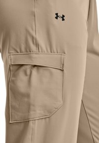 Béžové kalhoty Under Armour s velkou obdélníkovou kapsou se klopou na stehně a černým logem značky nad ní.