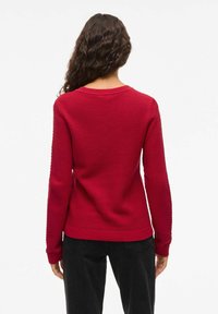 Pull en maille texturée rouge avec un col rond, des manches longues, et des poignets et un ourlet côtelés. La vue de dos montre un motif en nid d'abeille et une coupe décontractée.