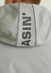Svijetlo siva jakna s kapuljačom od glatkog materijala, s hrabrim crnim natpisom "ASIN'" na leđima i kontrastnim detaljima šivanja.