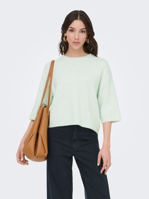 Jonge vrouw die een lichtmintgroene trui en zwarte broek draagt, met een grote beige leren schoudertas tegen een witte achtergrond.