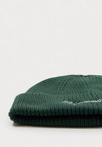 Green knitted beanie with ribbed texture, featuring a folded cuff and embroidered detail in white thread. Compact and close-up view.
