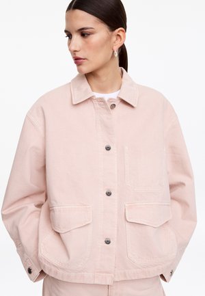 Kobieta w jasno różowej, oversize'owej kurtce zapinanej na guziki, z przednimi klapkowymi kieszeniami, patrząca w lewo na białym tle.