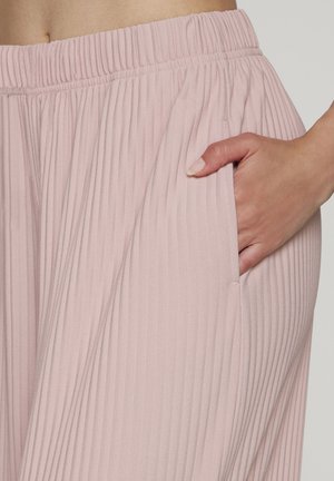 Hand in eine Seitentasche eines hellrosa, plissierten Rocks mit elastischem Bund auf neutralem Hintergrund gesteckt.