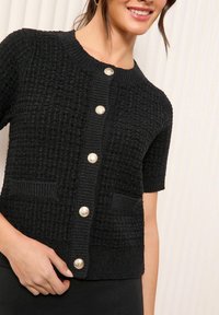 Melna adīta jaka ar īsām piedurknēm, kurai ir strukturēts audums, noapaļota dekoltē un zeltainas pogas, kā arī divas priekšējās kabatas.